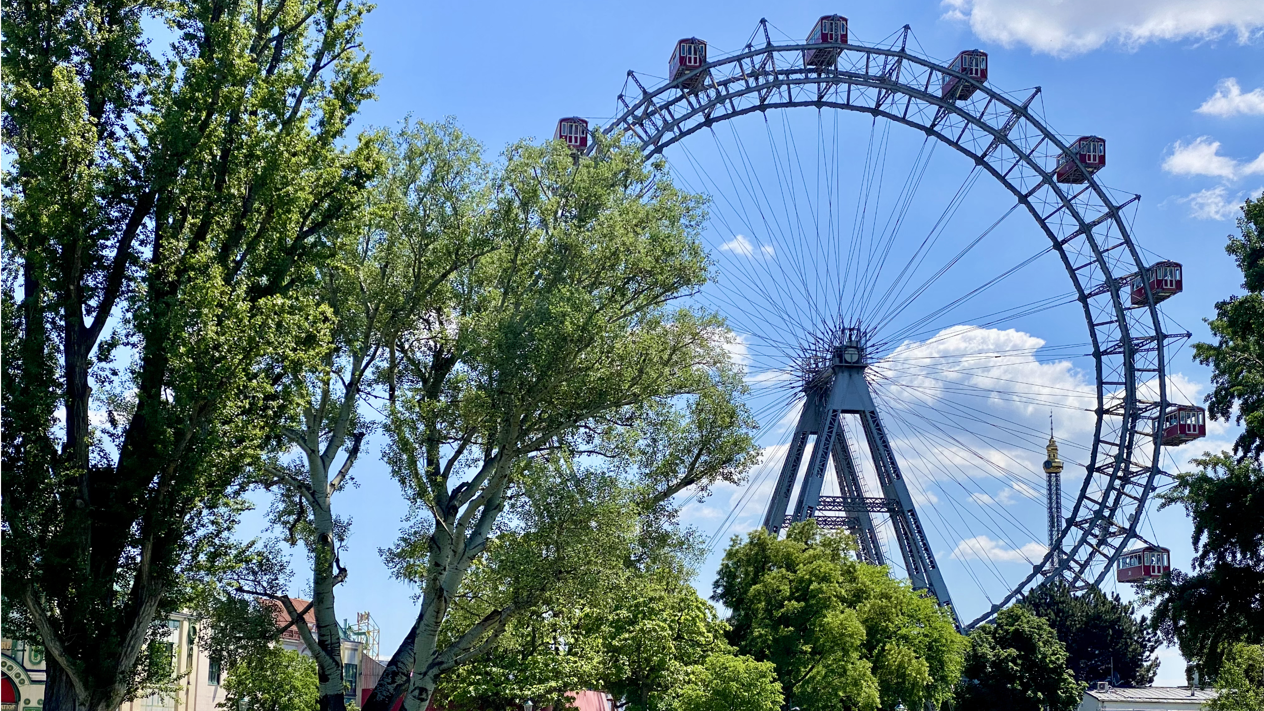 Das Wiener Riesenrad vor blauem Himmel, leicht bewölkt, grüne Bäume im Vordergrund.