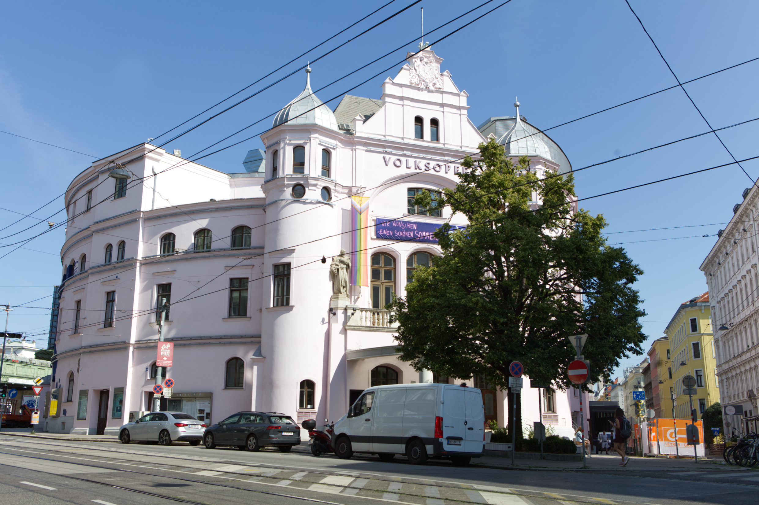 Die Wiener Volksoper in der Währinger Straße von außen. Ein großes weißes Gebäude mit zwei kleinen Türmen links und rechts des Portals. Die Rückseite ist zu einer Rundung geformt. Vor dem Gebäude steht ein Baum.