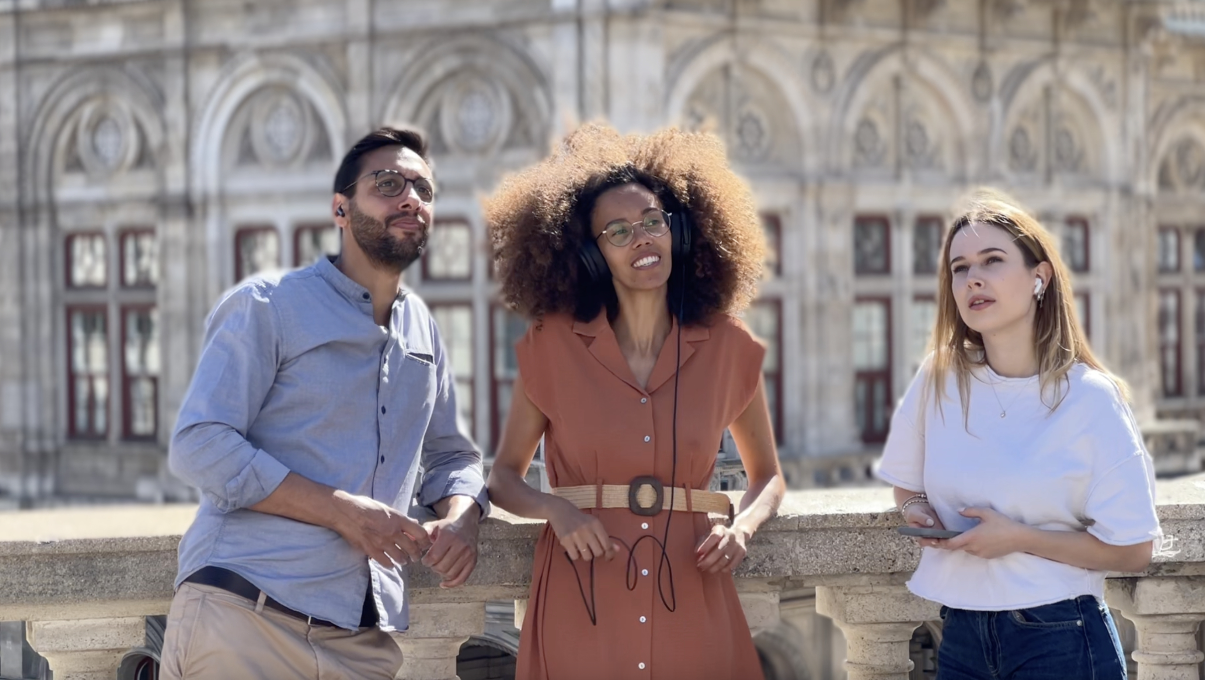 A man and two women are leaning against a balustrade, listening to the FILMED ON LOCATION audio tour through headphones. In the background, the Vienna State Opera is visible. It is a sunny summer day.