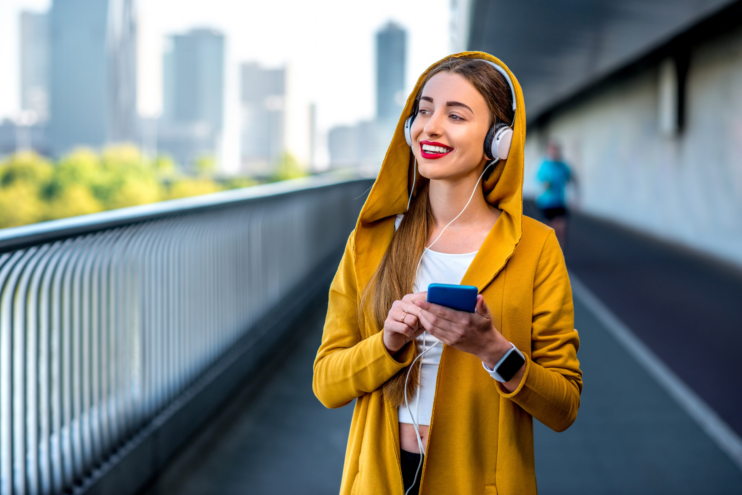 Junge Frau in einem gelben Mantel hört sich die FILMED ON LOCATION Audiotour an, mit einem Smartphone in der Hand auf der Wiener Reichsbrücke. Im Hintergrund sind Hochhäuser zu sehen.